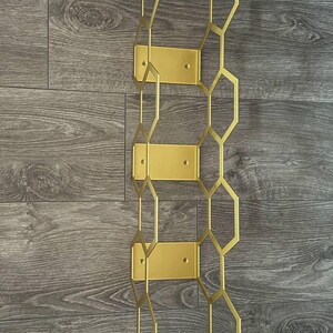 Puede incluir: Un botellero de metal dorado con un dise&ntilde;o geom&eacute;trico de panal. El botellero est&aacute; montado sobre una superficie de madera. El botellero tiene cuatro placas de montaje rectangulares y una serie de formas hexagonales interconectadas.