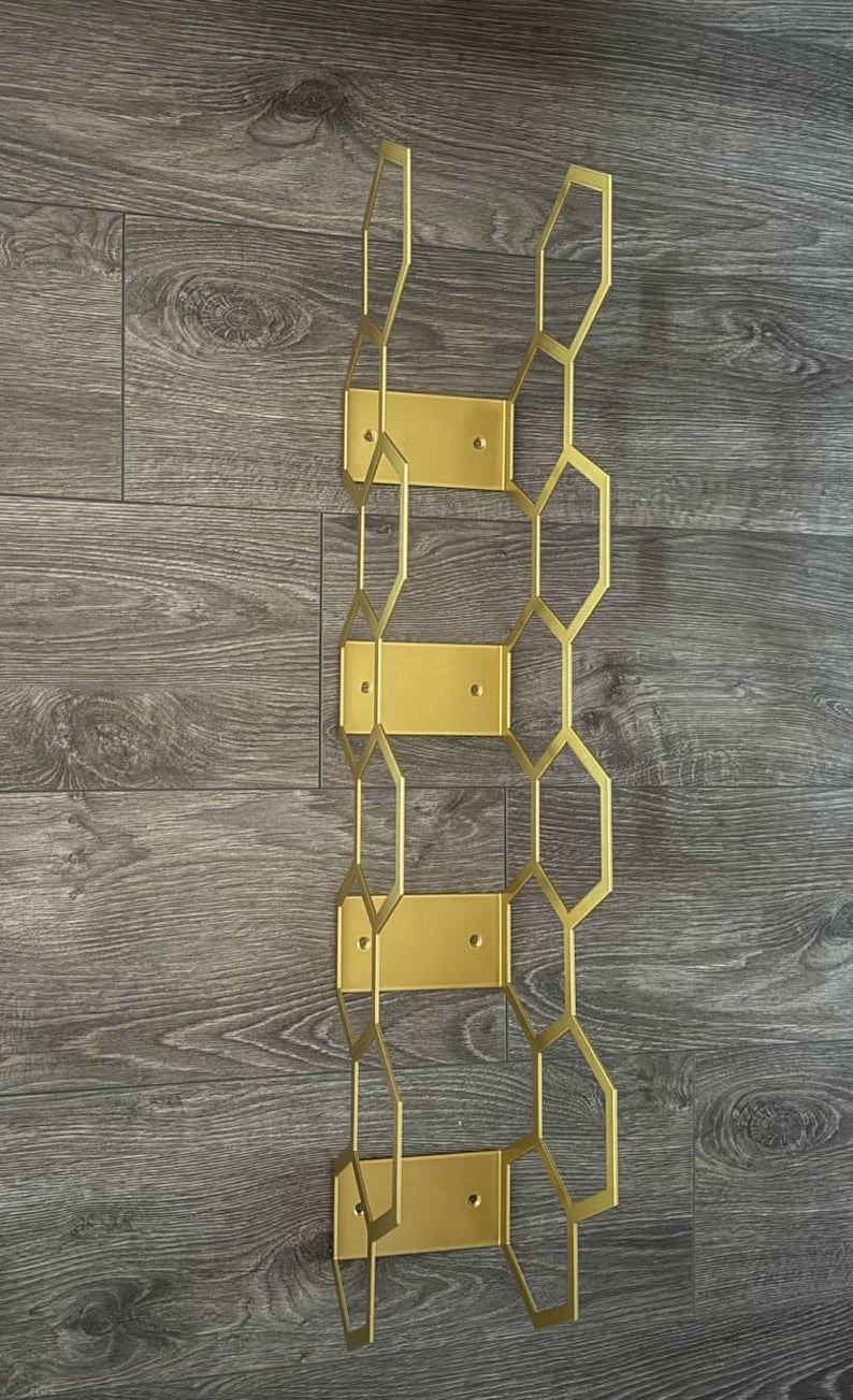 Puede incluir: Un botellero de metal dorado con un dise&ntilde;o geom&eacute;trico de panal. El botellero est&aacute; montado sobre una superficie de madera. El botellero tiene cuatro placas de montaje rectangulares y una serie de formas hexagonales interconectadas.