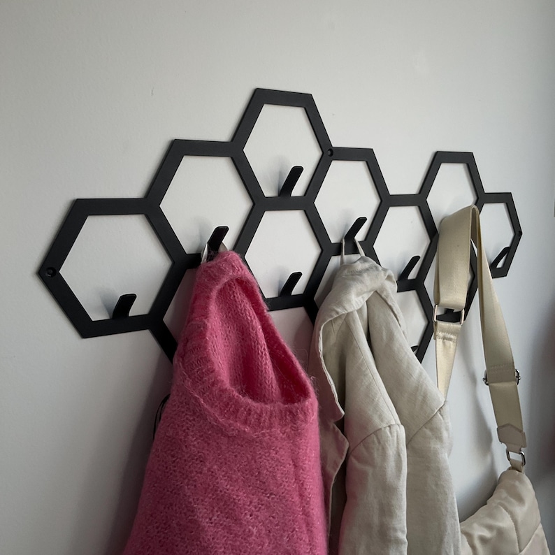 May include: Black metal wall-mounted coat rack in a honeycomb design. The rack holds a pink sweater, a beige jacket, and a cream-colored bag. The rack is mounted on a white wall.