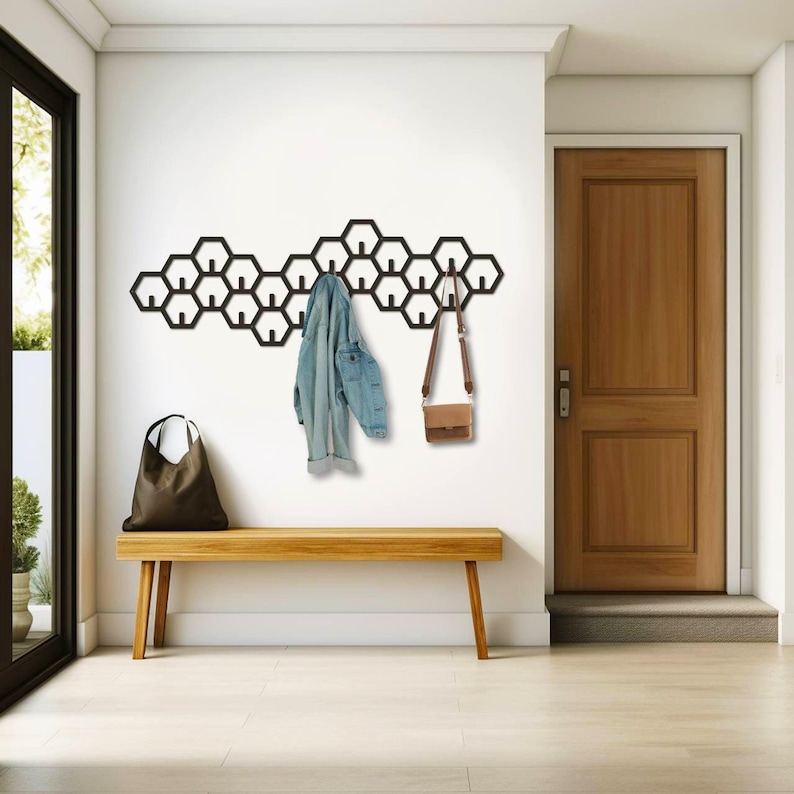 May include: A modern entryway featuring a black honeycomb wall-mounted coat rack. A light blue denim jacket and a brown leather purse hang from the rack. A wooden bench and a brown tote bag are below the rack, with a wooden door and a window in the background.