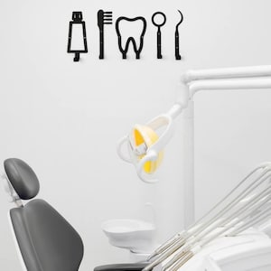 Peut inclure: Un ensemble de cinq silhouettes murales en m&eacute;tal noir repr&eacute;sentant des outils dentaires et une dent. Les ic&ocirc;nes incluent un tube de dentifrice, une brosse &agrave; dents, une molaire, un miroir dentaire et une sonde, dispos&eacute;es horizontalement.