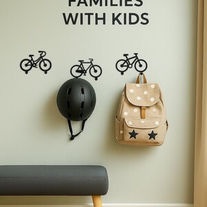 Op de afbeelding: Een wanddisplay met zwarte fietshaken, een zwarte helm en een beige rugzak met witte stippen en sterren. De tekst "PERFECT FOR FAMILIES WITH KIDS" staat boven de items.
