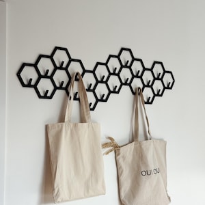 May include: A black metal honeycomb wall hook with multiple hooks, holding two beige tote bags. One bag has the text "OUI OUI" printed on it. A sprig of wheat is tucked into one of the bags.