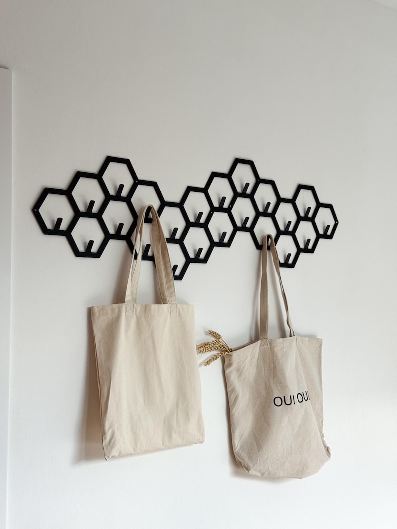 May include: A black metal honeycomb wall hook with multiple hooks, holding two beige tote bags. One bag has the text "OUI OUI" printed on it. A sprig of wheat is tucked into one of the bags.