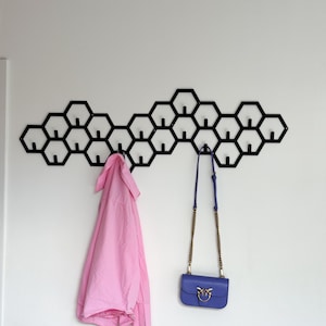 May include: A black metal wall-mounted coat rack with a honeycomb design. A pink shirt and a blue purse with a gold chain hang from the hooks. The rack is mounted on a white wall.