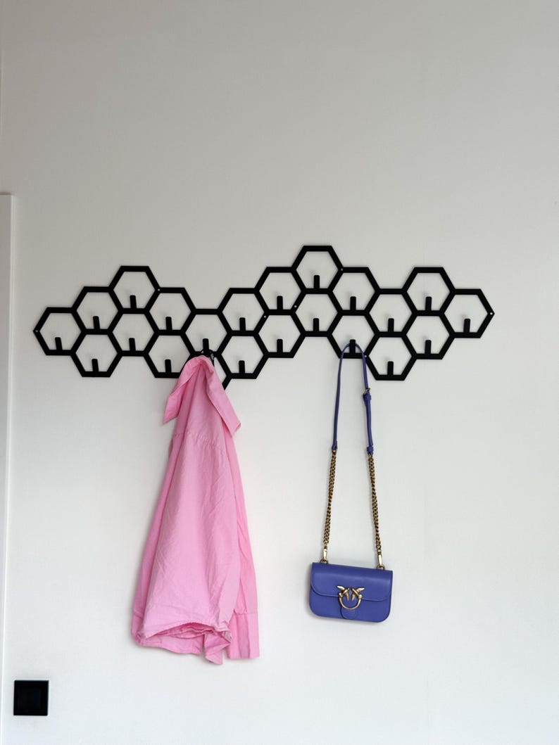 May include: A black metal wall-mounted coat rack with a honeycomb design. A pink shirt and a blue purse with a gold chain hang from the hooks. The rack is mounted on a white wall.