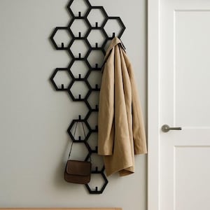 Może przedstawiać: Czarny, ścienny wieszak na płaszcze w kształcie plastra miodu z wieloma haczykami. Beżowy płaszcz i brązowa torebka wiszą na wieszaku. Wieszak jest zamontowany na jasnoszarej ścianie obok białych drzwi i drewnianej ławki.