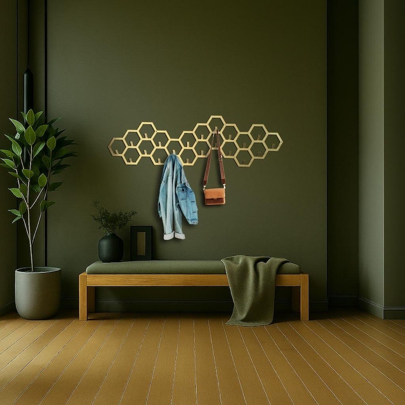 May include: A gold-colored, honeycomb-shaped wall-mounted coat rack with multiple hooks. A denim jacket and a brown purse hang from the rack. The rack is mounted on a dark green wall above a wooden bench with a green cushion and blanket.