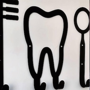 Peut inclure: Un ensemble de pat&egrave;res murales en m&eacute;tal noir comprenant un motif central en forme de dent avec deux crochets inf&eacute;rieurs, flanqu&eacute; d'un crochet en forme de brosse &agrave; dents &agrave; gauche et d'un miroir dentaire &agrave; droite, sur fond blanc.