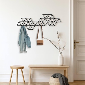 以下が含まれることがあります： デニムジャケットと茶色のハンドバッグが掛けられた、黒い幾何学模様の壁掛けコートラック。木製のベンチ、スツール、枝の入った花瓶も画像にあります。部屋はニュートラルなカラーパレットです。