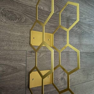 Puede incluir: Decoraci&oacute;n de pared de metal dorado con dise&ntilde;o de panal. El patr&oacute;n geom&eacute;trico est&aacute; compuesto por hex&aacute;gonos interconectados. La decoraci&oacute;n est&aacute; montada sobre placas rectangulares doradas, con orificios para tornillos visibles.