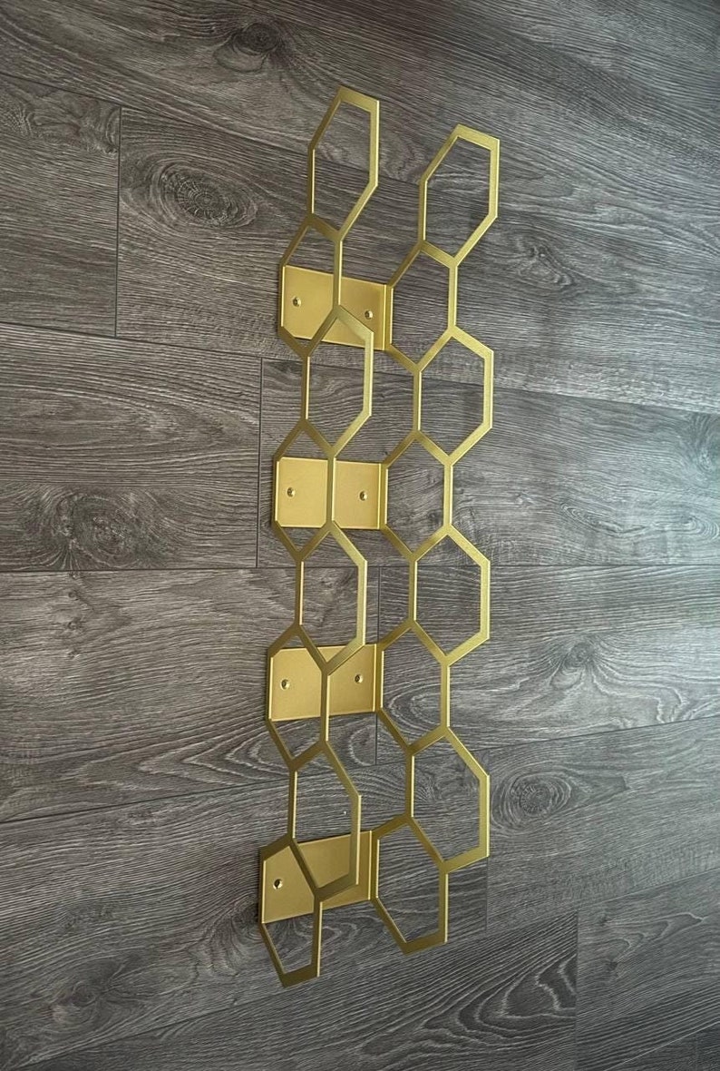 Puede incluir: Una pieza de decoraci&oacute;n mural geom&eacute;trica dorada, con un dise&ntilde;o de panal de abeja. La estructura met&aacute;lica est&aacute; compuesta por hex&aacute;gonos interconectados y placas de montaje rectangulares. La decoraci&oacute;n est&aacute; montada sobre una superficie de madera.