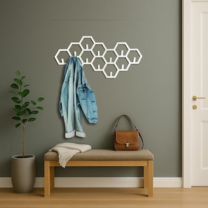 May include: A white honeycomb-shaped wall-mounted coat rack with multiple hooks. A light blue denim jacket hangs from the rack. A brown leather purse sits on a bench with a beige throw. A potted plant is on the left.