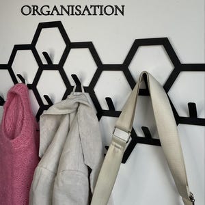 May include: Black metal wall-mounted coat rack with a honeycomb design, holding a pink sweater, a light-colored jacket, and a cream-colored bag. The text "12 HAKEN F&Uuml;R MAXIMALE ORGANISATION" is above the rack.