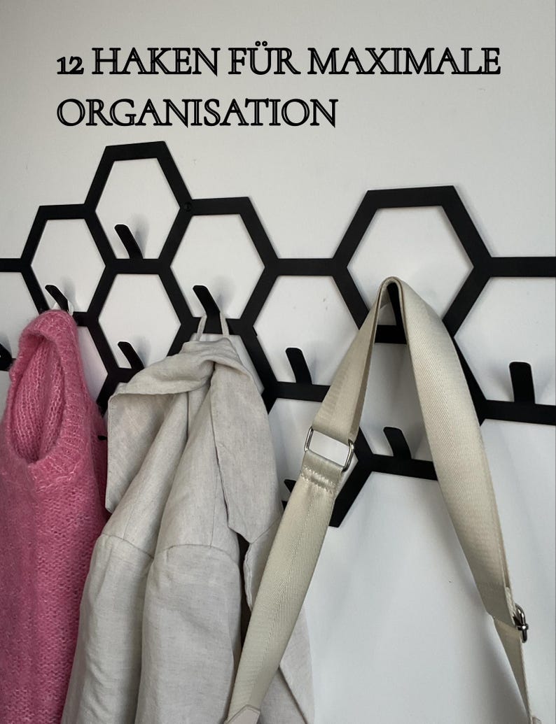 May include: Black metal wall-mounted coat rack with a honeycomb design, holding a pink sweater, a light-colored jacket, and a cream-colored bag. The text "12 HAKEN F&Uuml;R MAXIMALE ORGANISATION" is above the rack.