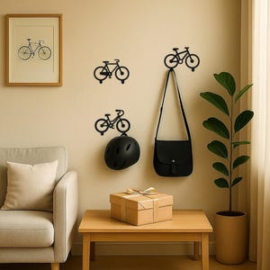 Op de afbeelding: Een kamer met fietsvormige wandhaken, een ingelijste fietsafdruk en een kleine houten tafel. Een zwarte helm en tas hangen aan de haken. Een cadeauverpakking met een gouden lint staat op de tafel. Een plant in een pot staat rechts.