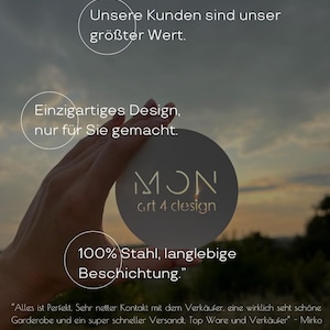 May include: A round, metallic object with the text "MON art 4 clesign" is held up against a blurred outdoor background. The object has the text "100% Stahl, langlebige Beschichtung." and other German text in a circle.