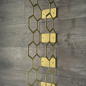 Puede incluir: Un botellero de pared de metal dorado con un dise&ntilde;o geom&eacute;trico de panal. El botellero est&aacute; compuesto por formas hexagonales interconectadas y placas de montaje rectangulares, creando una soluci&oacute;n de almacenamiento moderna y elegante. Est&aacute; montado en una pared de madera gris.