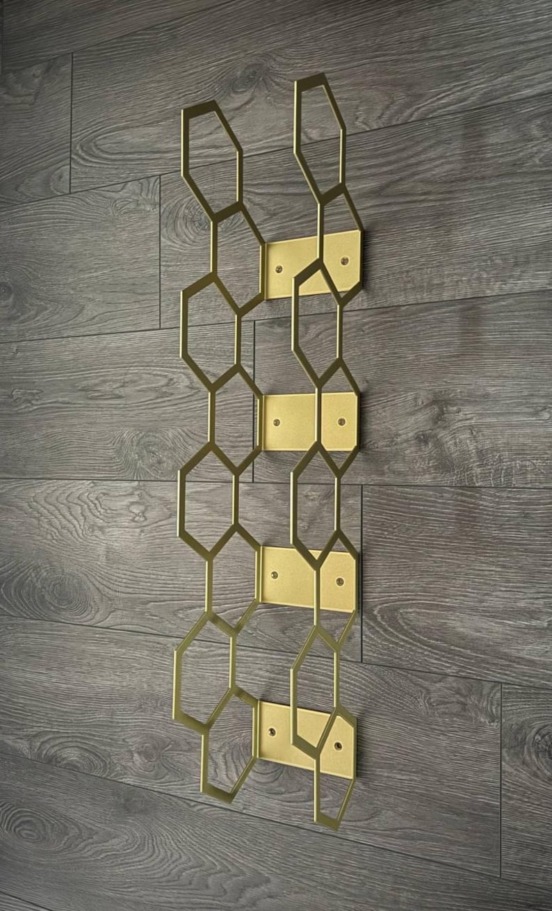 Puede incluir: Un botellero de pared de metal dorado con un dise&ntilde;o geom&eacute;trico de panal. El botellero est&aacute; compuesto por formas hexagonales interconectadas y placas de montaje rectangulares, creando una soluci&oacute;n de almacenamiento moderna y elegante. Est&aacute; montado en una pared de madera gris.