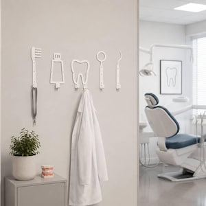 Op de afbeelding: Een set van vijf witte wandhaken in de vorm van tandheelkundige instrumenten, waaronder een tandenborstel, tandpastatube, tand, spiegel en een sonde. Een witte doktersjas hangt aan de tandhaak en een grijs koord hangt aan de tandenborstelhaak.