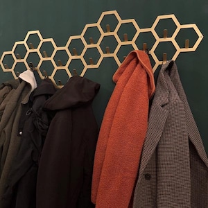 May include: A gold-colored honeycomb-shaped wall-mounted coat rack with multiple hooks. Various coats and jackets in shades of black, gray, and orange hang from the hooks, against a dark green wall. The rack adds a decorative and functional element.