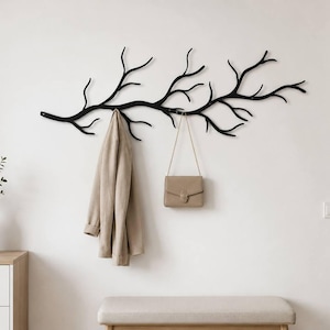 May include: A matte black wall-mounted coat rack shaped like a sprawling tree branch with multiple hooks. A beige jacket hangs from one of the lower branches, and a small beige shoulder bag with a thin gold chain strap hangs from another branch.