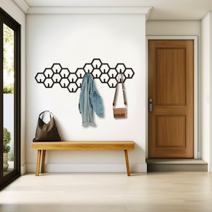 Könnte beinhalten: Ein schwarzer, wabenförmiger Wand-Kleiderhaken mit Haken. Eine Jeansjacke und eine braune Handtasche hängen an den Haken. Eine Holzbank steht vor dem Kleiderhaken.