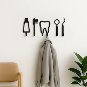 Op de afbeelding: Een set van vijf zwarte wandhaken in de vorm van tandheelkundige instrumenten, waaronder een tube tandpasta, een tandenborstel, een tand, een spiegel en een sonde. Een lichtgrijze blazer hangt aan de middelste tandvormige haak aan de muur.