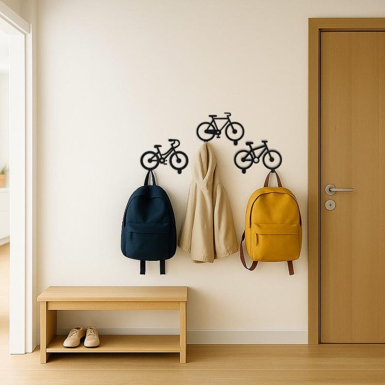 Op de afbeelding: Een gang met drie zwarte fietsmuurhaken. Een marineblauwe rugzak, een beige jas en een gele rugzak hangen aan de haken. Onder de haken staat een houten bank met twee paar schoenen.
