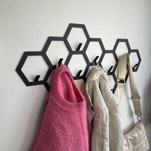 Könnte beinhalten: Schwarzer, wabenförmiger Wand-Kleiderständer mit zwei rosafarbenen Pullovern und einer braunen und weißen Tasche, die daran hängen.