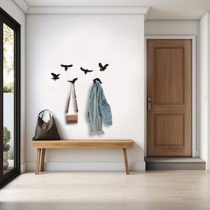 Op de afbeelding: Een lichtbruine houten bank staat onder een witte muur versierd met zwarte vogel silhouetten. Een bruine tote bag, een kleine crossbody tas en een spijkerjas hangen aan de muur. Een houten deur is zichtbaar op de achtergrond.