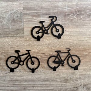 Op de afbeelding: Drie zwarte metalen wandhaken in de vorm van een fiets. Elke haak heeft een fiets silhouet met een haak onder het frame. De haken zijn gemonteerd op een lichtbruin houten oppervlak.