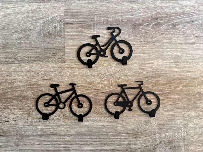 Op de afbeelding: Drie zwarte metalen wandhaken in de vorm van een fiets. Elke haak heeft een fiets silhouet met een haak onder het frame. De haken zijn gemonteerd op een lichtbruin houten oppervlak.
