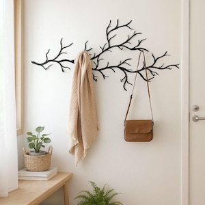 Peut inclure: Un porte-manteau mural en métal noir en forme de branche d'arbre avec plusieurs crochets. Une écharpe en tissu beige et un sac à bandoulière en cuir marron sont suspendus aux branches. Le porte-manteau est fixé sur un mur blanc uni.
