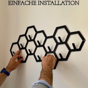 May include: Black metal wall-mounted coat rack in a honeycomb design. The rack features multiple hexagonal shapes with integrated hooks for hanging items. The text "EINFACHE INSTALLATION" is displayed above the rack.