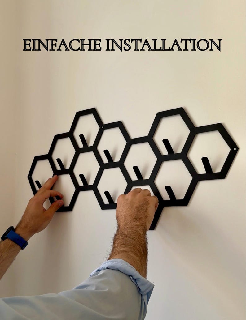 May include: Black metal wall-mounted coat rack in a honeycomb design. The rack features multiple hexagonal shapes with integrated hooks for hanging items. The text "EINFACHE INSTALLATION" is displayed above the rack.