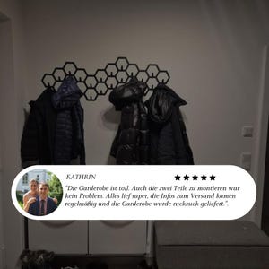 May include: A black honeycomb-shaped wall-mounted coat rack with several black jackets hanging on it. The rack is mounted on a white wall. A customer review is visible at the bottom of the image.