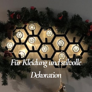 May include: A black metal honeycomb-shaped wall decoration with snowflake-shaped lights. The decoration is surrounded by a garland of green pine branches, red berries, and pinecones. Text on the image reads "F&uuml;r Kleidung und stilvolle Dekoration."
