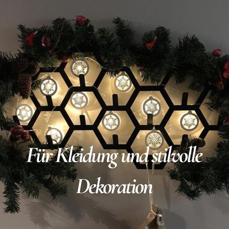 May include: A black metal honeycomb-shaped wall decoration with snowflake-shaped lights. The decoration is surrounded by a garland of green pine branches, red berries, and pinecones. Text on the image reads "F&uuml;r Kleidung und stilvolle Dekoration."