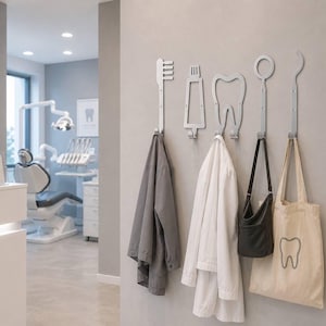 Op de afbeelding: Een set van vijf zilverkleurige wandhaken in de vorm van tandheelkundige instrumenten, waaronder een tandenborstel, tandpastatube, tand, tandartsspiegel en een sonde. Elke haak is aan de muur bevestigd en houdt jassen en tassen vast.