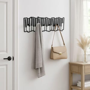 Puede incluir: Perchero de pared negro con forma de teclas de piano. Una bufanda gris y un bolso beige cuelgan del perchero. El perchero está montado en una pared blanca.