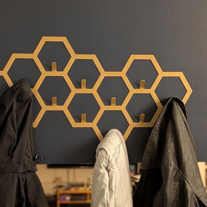 May include: A gold-colored honeycomb-shaped wall-mounted coat rack with multiple hooks. Three coats, including a black jacket, hang from the hooks. The rack is mounted on a dark blue wall, providing a modern and stylish storage solution.