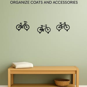 Op de afbeelding: Zwarte metalen fietsvormige haken aan een lichtgroene muur, met de tekst "UNIQUE & FUN BIKE HOOKS" en "ORGANIZE COATS AND ACCESSORIES". Daaronder een houten bank met een gevouwen cr&egrave;mekleurige deken en een rieten mand.
