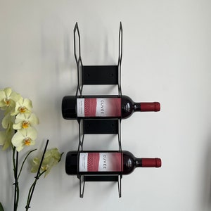 Puede incluir: Botellero de pared de metal negro que sostiene dos botellas de vino tinto. Las botellas de vino tienen etiquetas rojas con la palabra "CUV&Eacute;E" y tapones rojos. El botellero est&aacute; montado en una pared blanca.