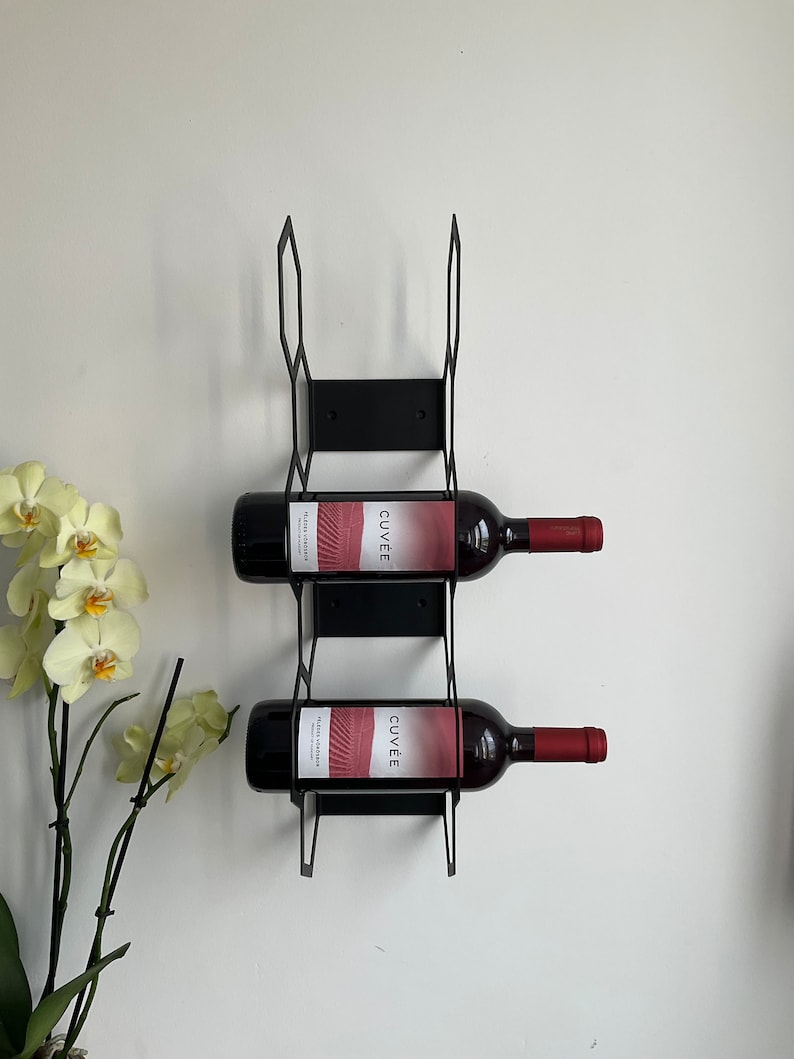 Puede incluir: Botellero de pared de metal negro que sostiene dos botellas de vino tinto. Las botellas de vino tienen etiquetas rojas con la palabra "CUV&Eacute;E" y tapones rojos. El botellero est&aacute; montado en una pared blanca.