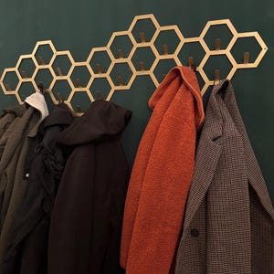 May include: A gold-colored metal wall-mounted coat rack featuring a geometric honeycomb pattern with multiple integrated hooks. Several winter coats in shades of black, olive green, burnt orange, and a brown checkered pattern hang from the hooks.