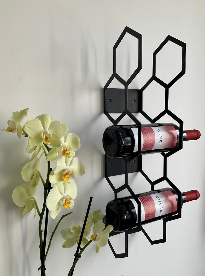 Puede incluir: Un botellero de metal negro con un dise&ntilde;o geom&eacute;trico de panal, que contiene dos botellas de vino tinto. Las botellas tienen etiquetas rojas y blancas. Un tallo de orqu&iacute;deas amarillas est&aacute; en primer plano, contra una pared blanca.