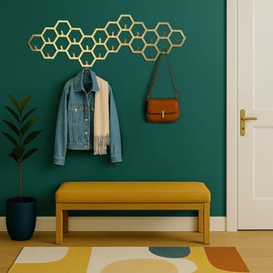 May include: A gold-colored, honeycomb-shaped wall-mounted coat rack with multiple hooks. A denim jacket and scarf hang from the rack, with a brown purse suspended from another hook. A yellow bench and a patterned rug complete the entryway.