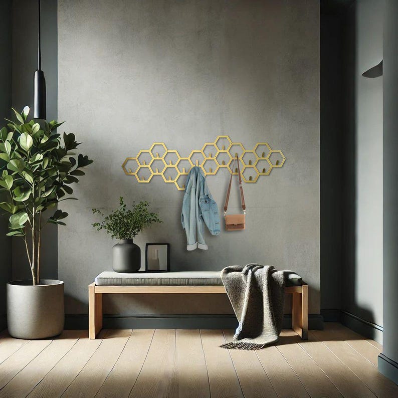 May include: A gold-colored, honeycomb-shaped wall-mounted coat rack with multiple hooks. A denim jacket and a small brown purse hang from the rack. A bench with a gray cushion and a blanket are below the rack. A potted plant is on the left.