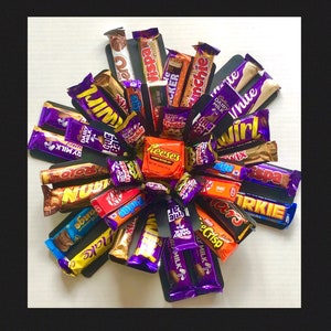 May include: A selection of chocolate bars, such as Dairy Milk, Wispa, and Kit Kat, arranged in a circular display. The wrappers are brightly coloured, with brand names clearly visible. A central cluster of Reese's peanut butter cups is also present.